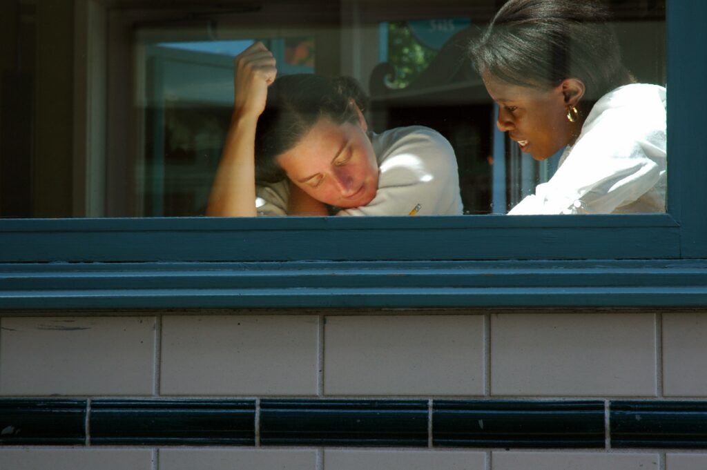 An image of two women through a window. One woman looking stressed while another talking while she writes