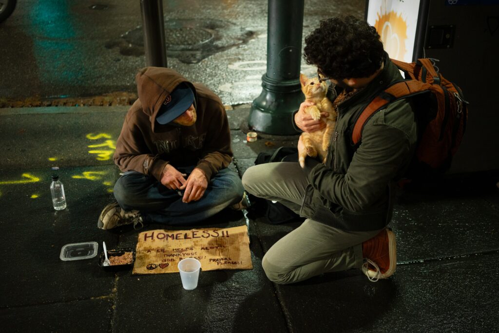 A man holding a kitten close in his arms kneeling next to a homeless man on the gorund