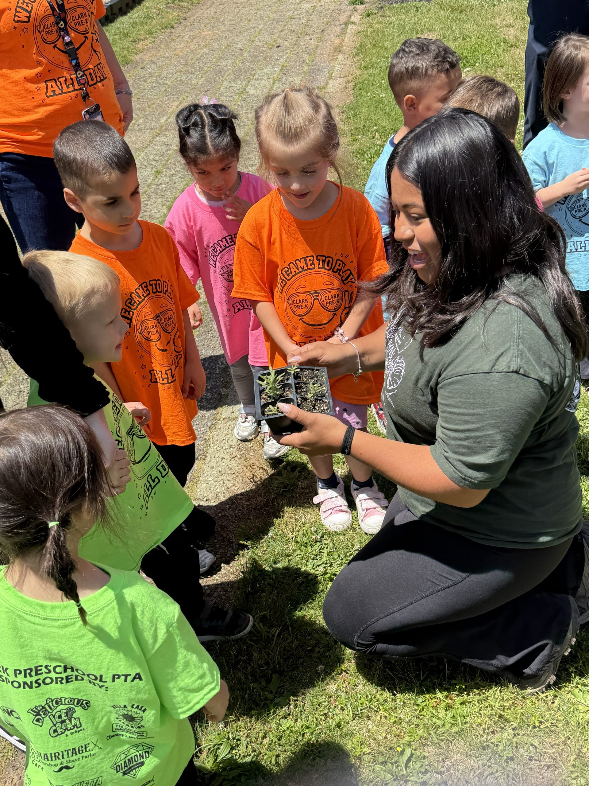 Close-up image of Clark Pre-K students observing a Groundworks Elizabeth staff member as she gives them instructions while holding a new plant