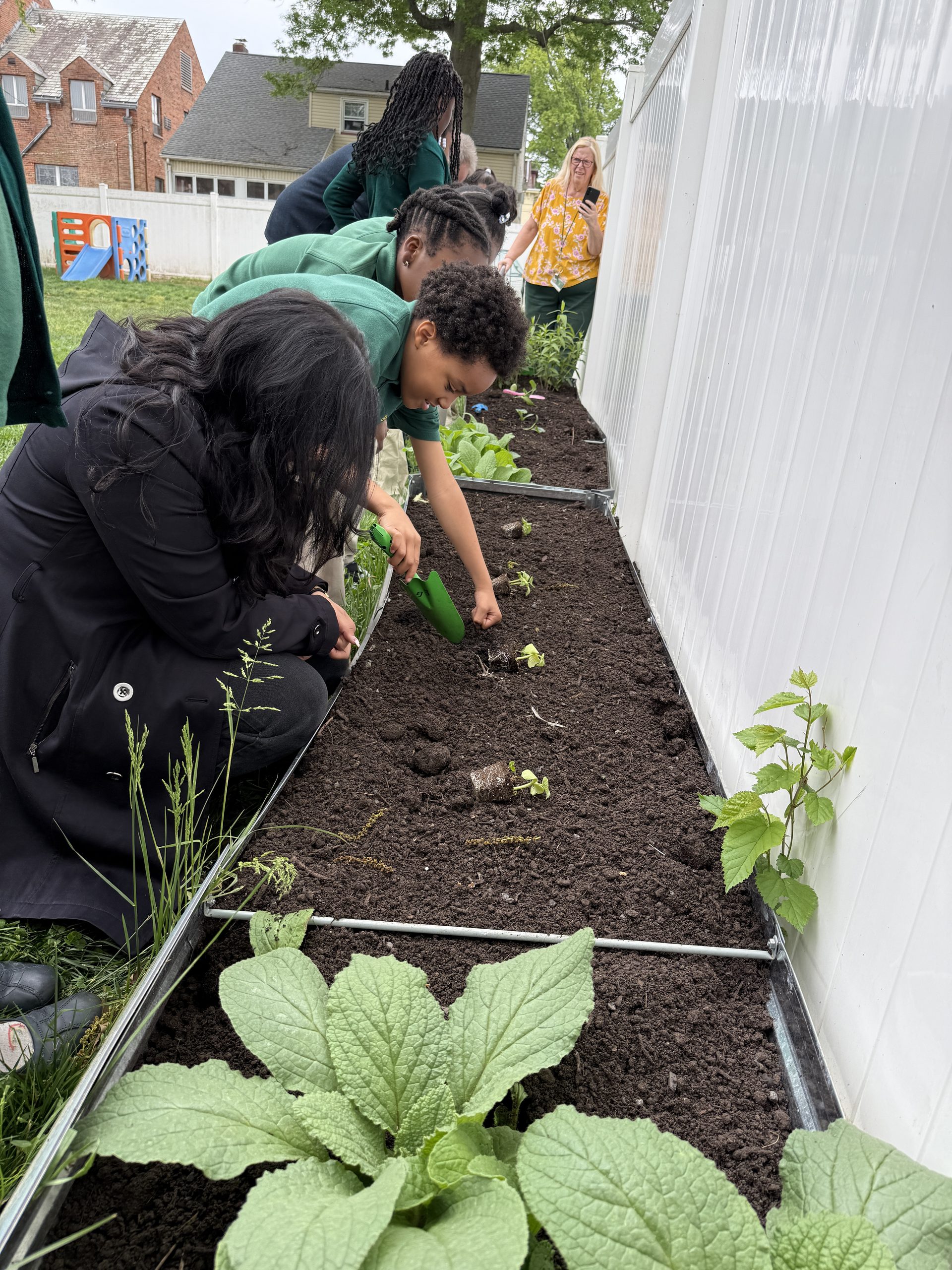 A group of students gets help from their teacher as they plant young seedlings in the raised garden bed