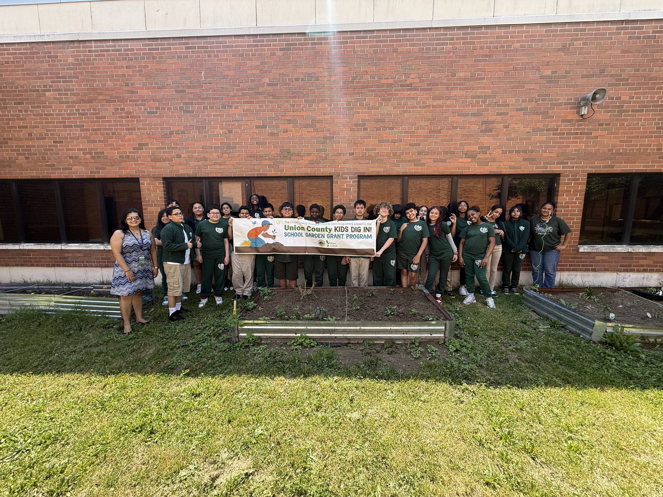 Jerome Dunn Academy students and Union County Representatives pose behind their raised garden beds, proudly displaying their Union County Kids Dig In School Garden Grant banner