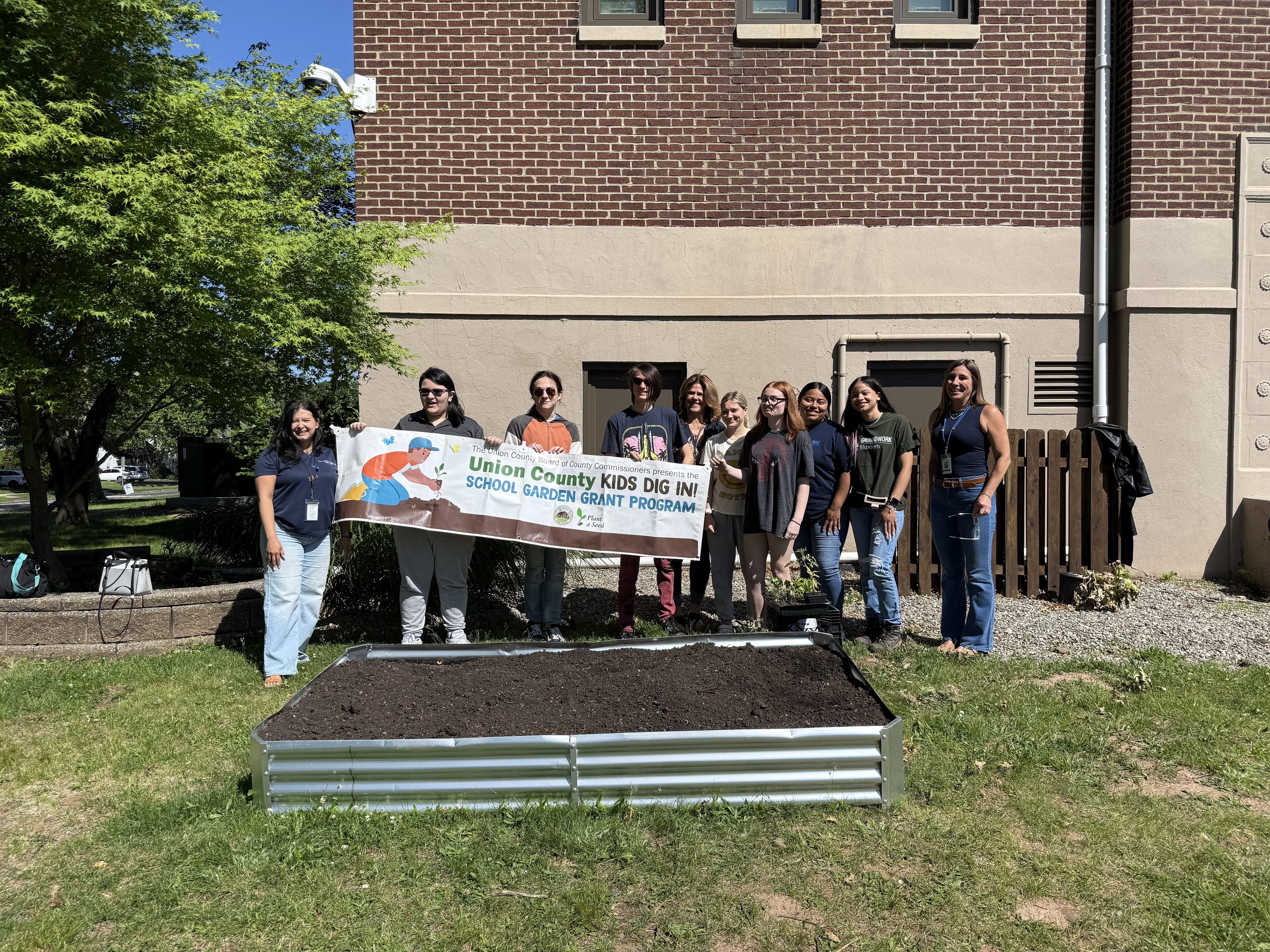 Lincoln School students, teachers and Groundworks Elizabeth representatives smiling for the camera while holding up their Kids Dig In Garden Grant Program banner