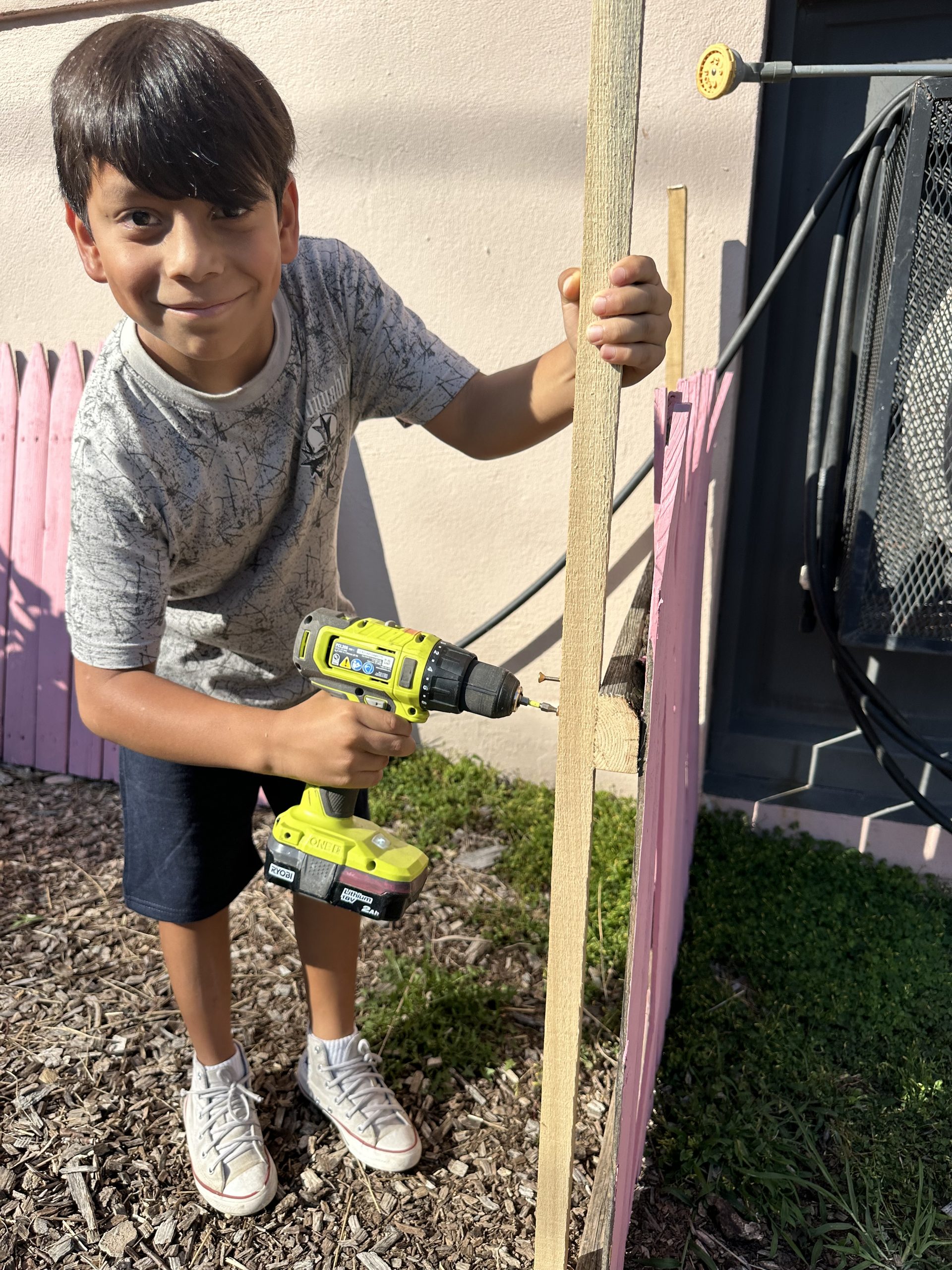 School No. 12 male student holding a green drill to drive a screw into the wooden pink fence