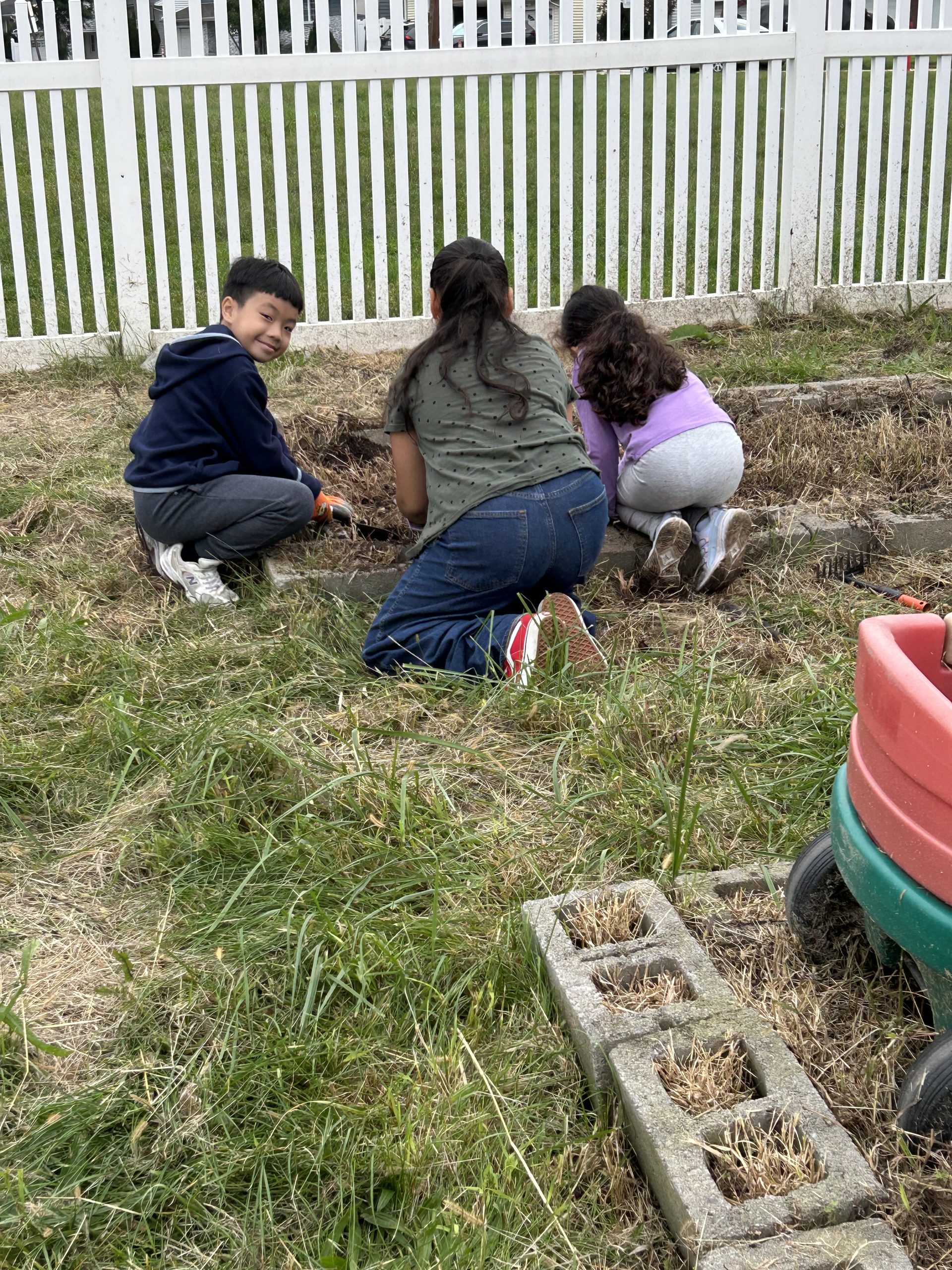 Image of three students kneeling on the ground as they clear the weeds out of a garden bed. One young student is smiling and facing the camera