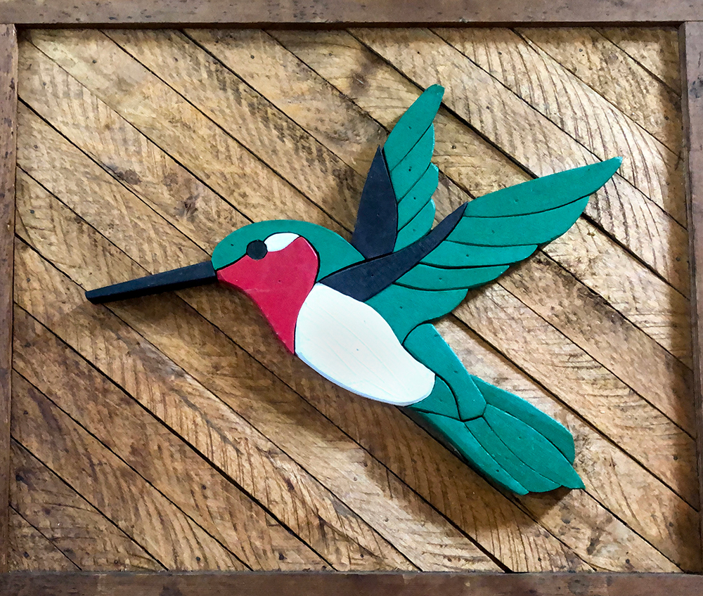 James Colangelo was awarded First Place for Craft by a Non-Professional artist for this work, titled “Ruby,” that depicts a colorful bird in flight. It was created using wood lathe, cedar, acrylic paint, a scroll saw, and a band saw. The bird is seen in profile with its wings spread and tilted upwards. The bird’s body is teal-blue, with a magenta blaze on its face, a white breast, and navy-blue edging on its wings. Its beak is very long and thin. The bird is mounted on natural-colored, unpainted cedar wood, creating a stark contrast with the bright paint used for the bird’s body. This piece has a pleasing simplicity.