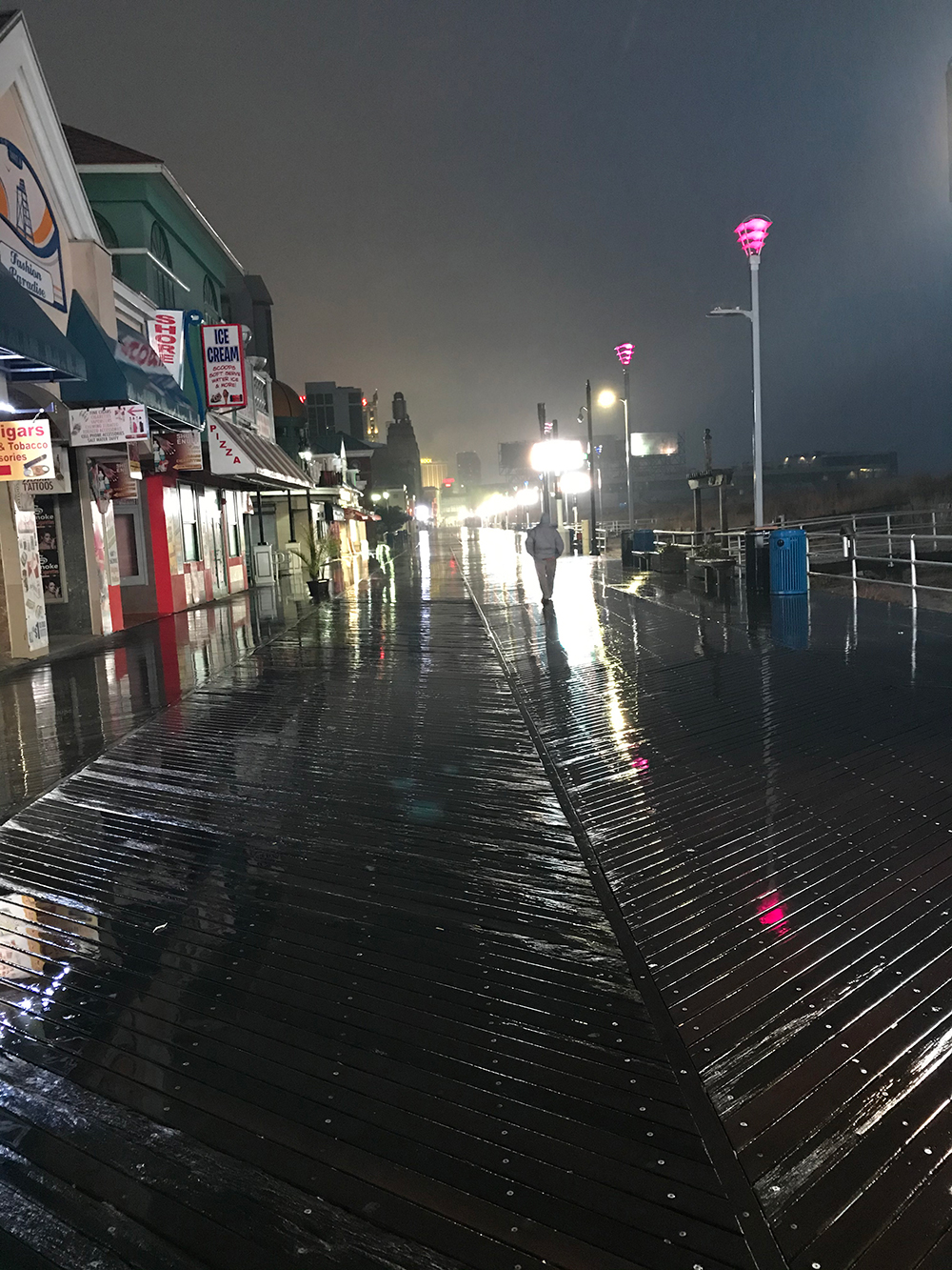 David Weisbrod was awarded First Place for Photography by a Non-Professional artist. Titled “Night Owl,” his photograph shows a nearly deserted boardwalk on a dark, rainy night. The boardwalk is almost devoid of people, except one man walking away from us, his profile made sharp by street lights just beyond him that cast a bright reflective glare on the wet boardwalk. Even if shadows in the distance are people, the lone man is the focus. The lower half of the composition is dark-grey, soaking-wet boardwalk planks. The overcast sky is dull and gray. On the left side, boardwalk businesses are closed for the night. Signs for pizza and ice cream. On the right, a ramp leads off, maybe towards the ocean, and a blurry hint of dune grass can be seen in the foggy darkness.